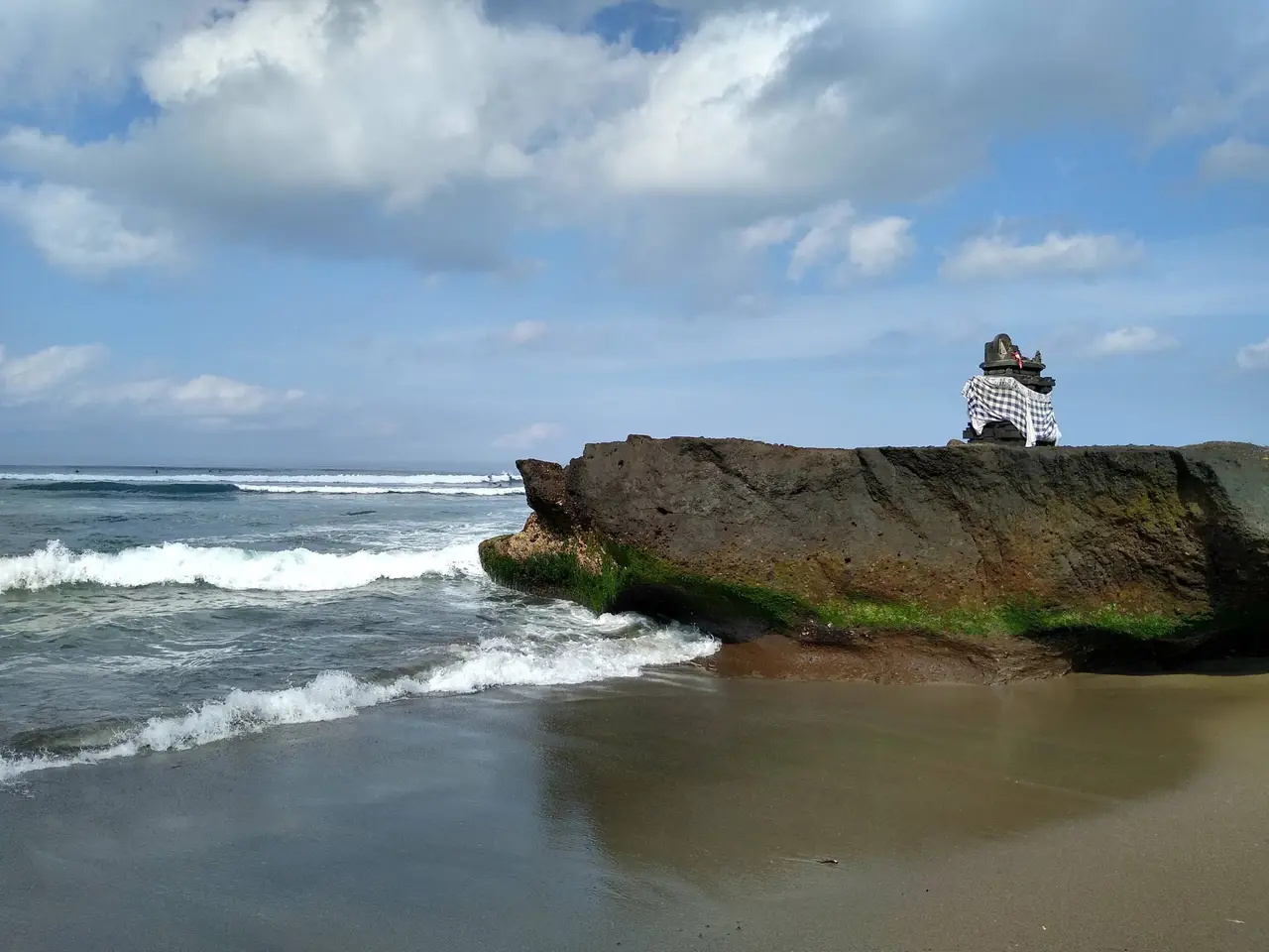 A dark rock with a small white-draped altar on top, standing in the surf on the beach at Canggu under a sky of broken cloud.