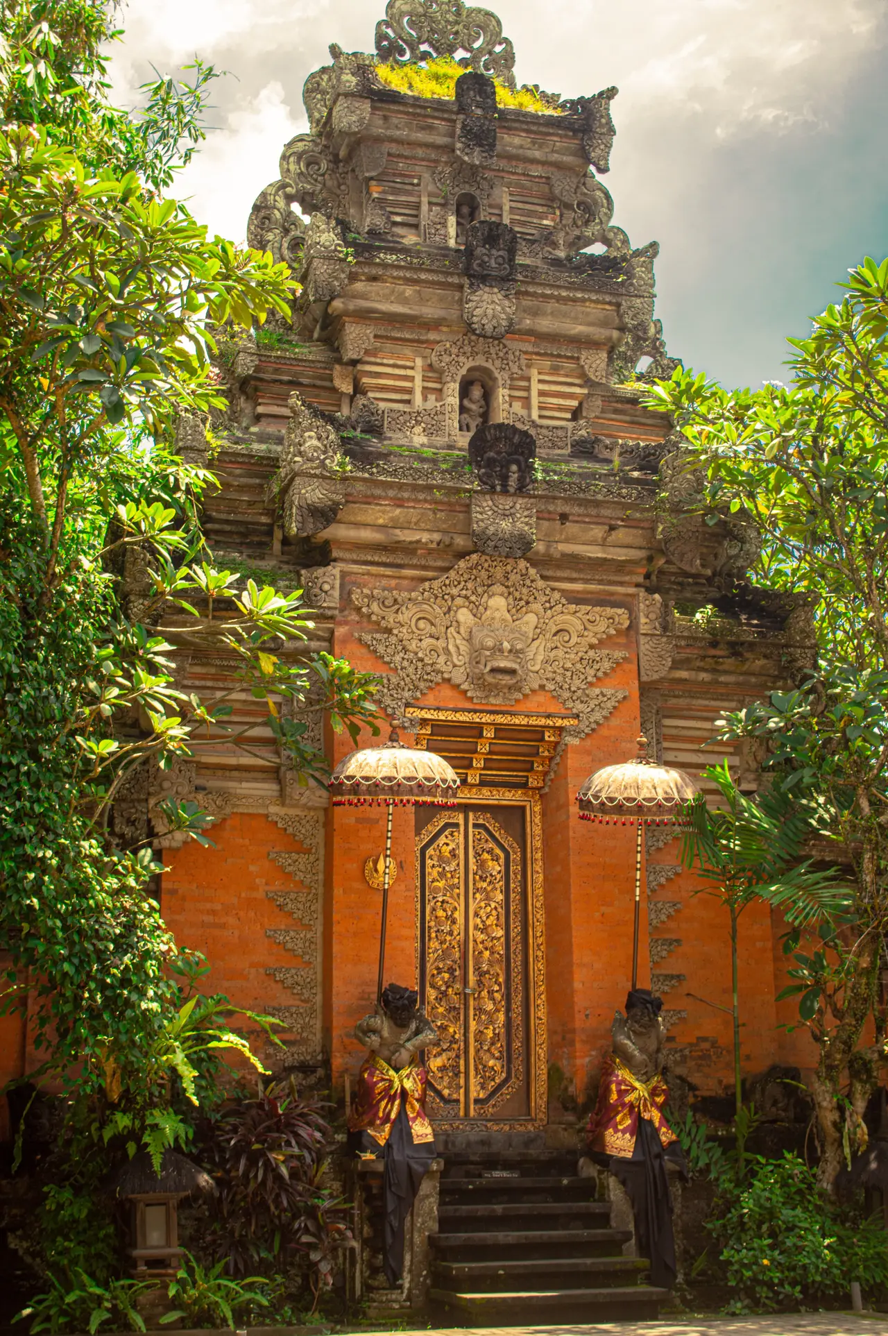 A carved stone gateway at Ubud Palace in orange brick and grey relief, with two small ceremonial umbrellas and green foliage framing it.
