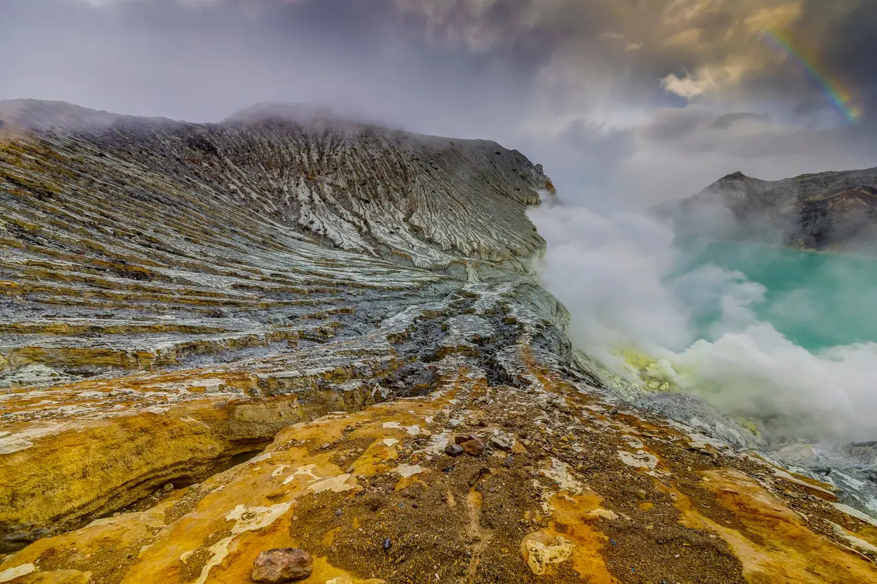 Sulphur-yellow rock and drifting steam on the rim of Kawah Ijen, with the turquoise crater lake beyond.