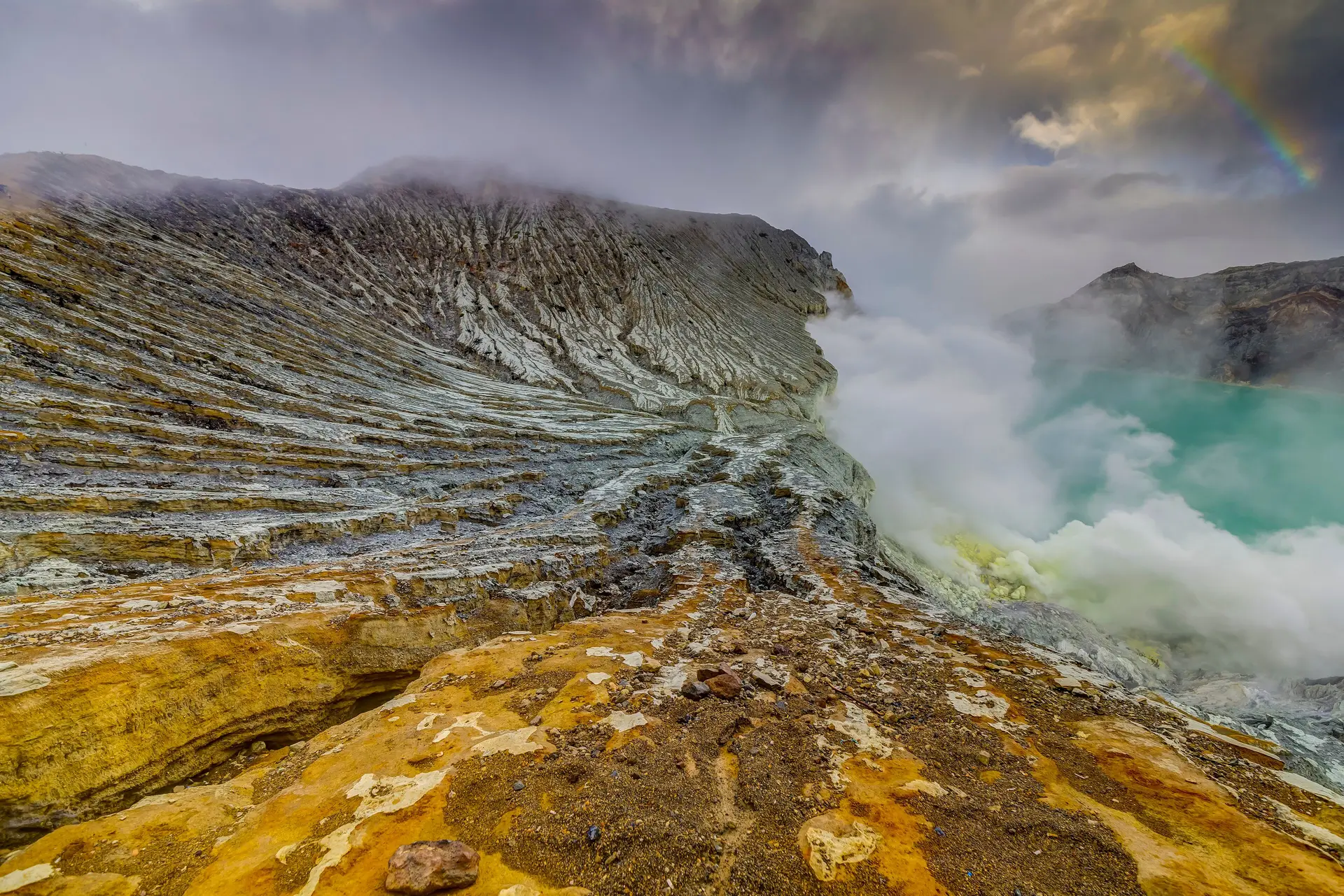 Sulphur-yellow rock and drifting steam on the rim of Kawah Ijen, with the turquoise crater lake beyond.