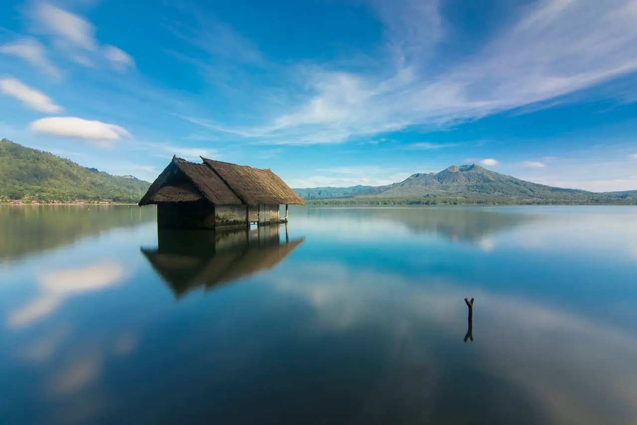 A thatched hut standing in still water on Lake Batur, mirrored in the lake, with Mount Batur low on the horizon under streaked blue sky.