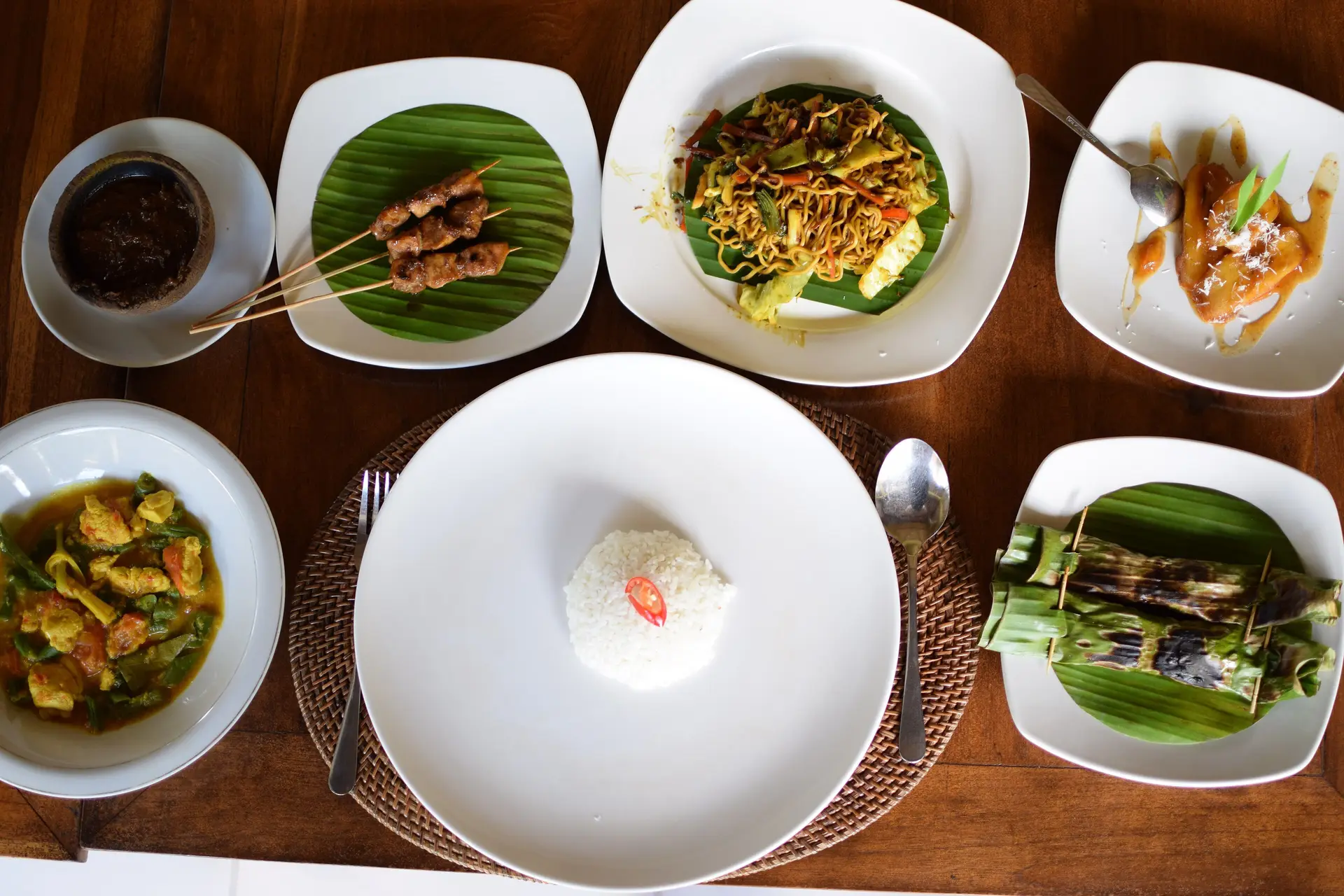 An overhead view of a Balinese meal: satay, rice, a banana-leaf parcel and small side dishes on white plates.