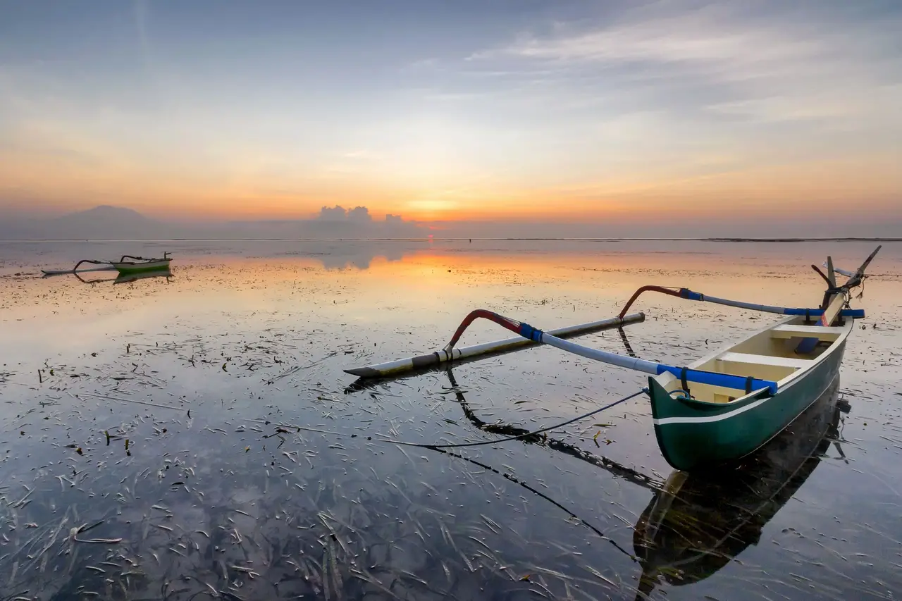 A green-and-white outrigger fishing boat (jukung) lying on the wet sand at low tide at Sanur, its reflection in the shallow water and the sky orange before sunrise.